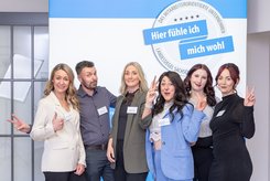 Sechs Personen stehen nebeneinander vor einem blau-weißen Messestand mit dem Schriftzug des Landessiegel „Hier fühle ich mich wohl“. 