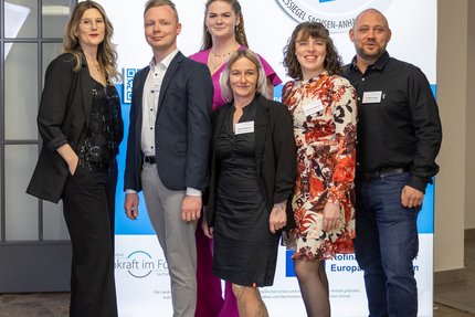 Sechs Personen stehen nebeneinander vor einem blau-weißen Messestand mit dem Schriftzug des Landessiegel „Hier fühle ich mich wohl“. 