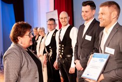 Urkundenübergabe auf einer Bühne: Eine Person überreicht einer anderen Person eine gerahmte Urkunde; daneben stehen mehrere weitere Teilnehmende in einer Reihe und blicken nach vorn.