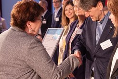 Übergabe einer Urkunde bei einer Veranstaltung: Zwei Personen stehen im Vordergrund und schütteln sich die Hände, während eine Urkunde überreicht wird; im Hintergrund stehen weitere Teilnehmende in einer Reihe auf der Bühne