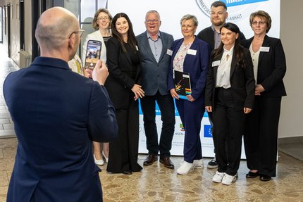Sieben Personen stehen nebeneinander vor einem blau-weißen Messestand mit dem Schriftzug des Landessiegel „Hier fühle ich mich wohl“. Eine Person fotografiert die sieben Menschen vor der Messewand.