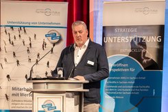 Redebeitrag bei einer Veranstaltung: Eine Person steht hinter einem Rednerpult und spricht in ein Mikrofon; hinter dem Podium ist ein Banner mit Begriffen wie Strategie, Unterstützung und Entwicklung zu sehen.