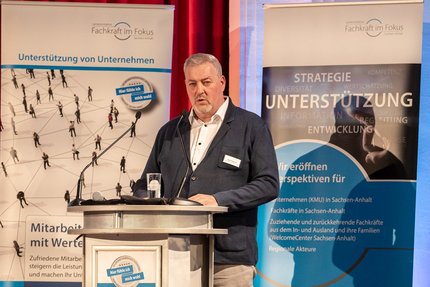 Redebeitrag bei einer Veranstaltung: Eine Person steht hinter einem Rednerpult und spricht in ein Mikrofon; hinter dem Podium ist ein Banner mit Begriffen wie Strategie, Unterstützung und Entwicklung zu sehen.