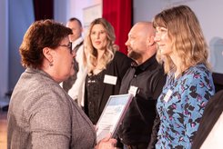 Urkundenübergabe auf einer Bühne: Eine Person überreicht einer anderen Person eine gerahmte Urkunde; daneben stehen mehrere weitere Teilnehmende in einer Reihe und blicken nach vorn.