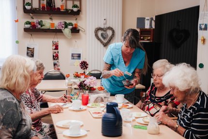vier ältere Frauen sitzen am Tisch und frühstücken. Eine jüngere Frau serviert Marmelade.
