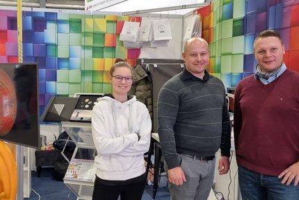 drei Menschen stehen auf einer Messe an ihrem Stand. Der Stand hat regenbogen-farbene Wände