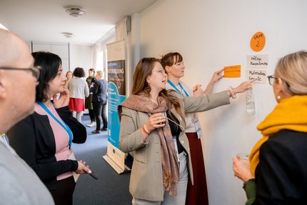 Eine Gruppe Menschen stehen an einer Wand und kleben beschriebene Moderationskarten an.