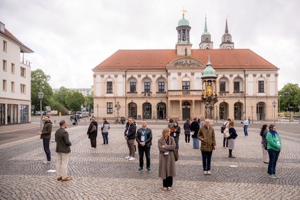 Mehrere Menschen stehen mit Abstand zueinander auf einem gepflasterten Platz vor einem historischen Gebäude mit Säulen, Rundbögen und rotem Ziegeldach. 