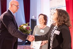 Zwei Personen stehen im Vordergrund und schütteln sich die Hände; Eine weitere Person steht daneben und schaut zu.