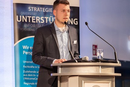 Redebeitrag bei einer Veranstaltung: Eine Person steht hinter einem Rednerpult und spricht in ein Mikrofon; hinter dem Podium ist ein Banner mit Begriffen wie Strategie, Unterstützung und Entwicklung zu sehen.
