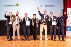 Gruppenfoto auf einer Bühne: Mehrere Teilnehmende stehen nebeneinander, halten Urkunden und Blumensträuße in den Händen und heben teilweise die Arme. Im Hintergrund ist eine Projektionsfläche mit der Überschrift „Herzlichen Glückwunsch!“ und Logos beteiligter Unternehmen zu sehen.