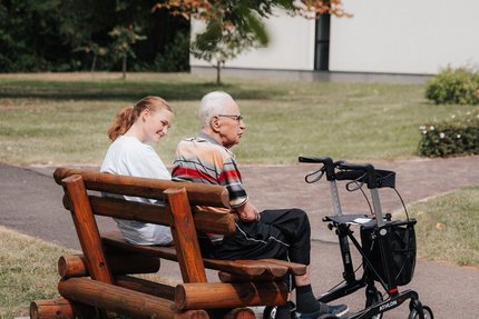 Eine junge Frau und ein alter Mann sitzen auf einer Bank in einer Grünanlage