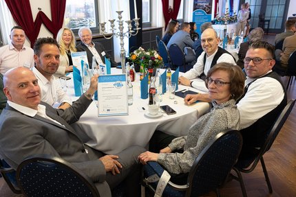 Mehrere Teilnehmende sitzen an einem runden Tisch in einem Veranstaltungssaal und blicken in die Kamera. Auf dem Tisch stehen Getränke, Gläser, Kerzenleuchter und eine gut sichtbare Urkunde mit der Aufschrift „Urkunde“.
