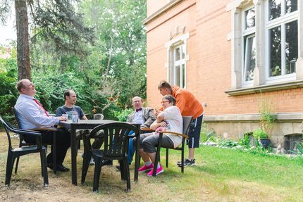 Eine Gruppe Menschen sitzen an einem Tisch auf einer Wiese vor einem Haus.
