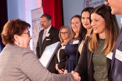 Übergabe einer Urkunde bei einer Veranstaltung: Zwei Personen stehen im Vordergrund und schütteln sich die Hände, während eine Urkunde überreicht wird; im Hintergrund stehen weitere Teilnehmende in einer Reihe auf der Bühne.