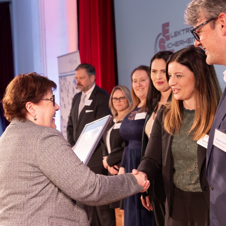 Übergabe einer Urkunde bei einer Veranstaltung: Zwei Personen stehen im Vordergrund und schütteln sich die Hände, während eine Urkunde überreicht wird; im Hintergrund stehen weitere Teilnehmende in einer Reihe auf der Bühne.
