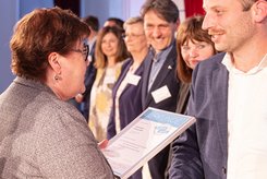 Übergabe einer Urkunde bei einer Veranstaltung: Zwei Personen stehen im Vordergrund und schütteln sich die Hände, während eine Urkunde überreicht wird; im Hintergrund stehen weitere Teilnehmende in einer Reihe auf der Bühne