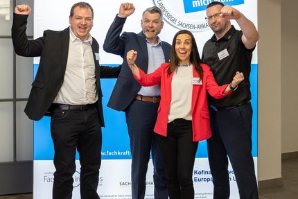 Vier Personen stehen nebeneinander vor einem blau-weißen Messestand mit dem Schriftzug des Landessiegel „Hier fühle ich mich wohl“. 