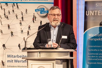 edebeitrag bei einer Veranstaltung: Eine Person steht hinter einem Rednerpult und spricht in ein Mikrofon; hinter dem Podium ist ein Banner mit Begriffen Unterstützung von Unternehmen