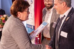 Übergabe einer Urkunde bei einer Veranstaltung: Zwei Personen stehen im Vordergrund und schütteln sich die Hände, während eine Urkunde überreicht wird; 