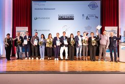 Gruppenfoto auf einer Bühne: Mehrere Teilnehmende stehen nebeneinander, halten Urkunden und Blumensträuße in den Händen und heben teilweise die Arme. Im Hintergrund ist eine Projektionsfläche mit der Überschrift „Herzlichen Glückwunsch!“ und Logos beteiligter Unternehmen zu sehen.