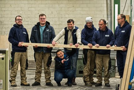 Eine Gruppe von Menschen stehen in einer Werkhalle und halten eine lange Holzlatte