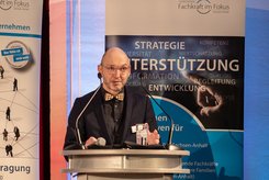 Redebeitrag bei einer Veranstaltung: Eine Person steht hinter einem Rednerpult und spricht in ein Mikrofon; hinter dem Podium ist ein Banner mit Begriffen wie Strategie, Unterstützung und Entwicklung zu sehen.