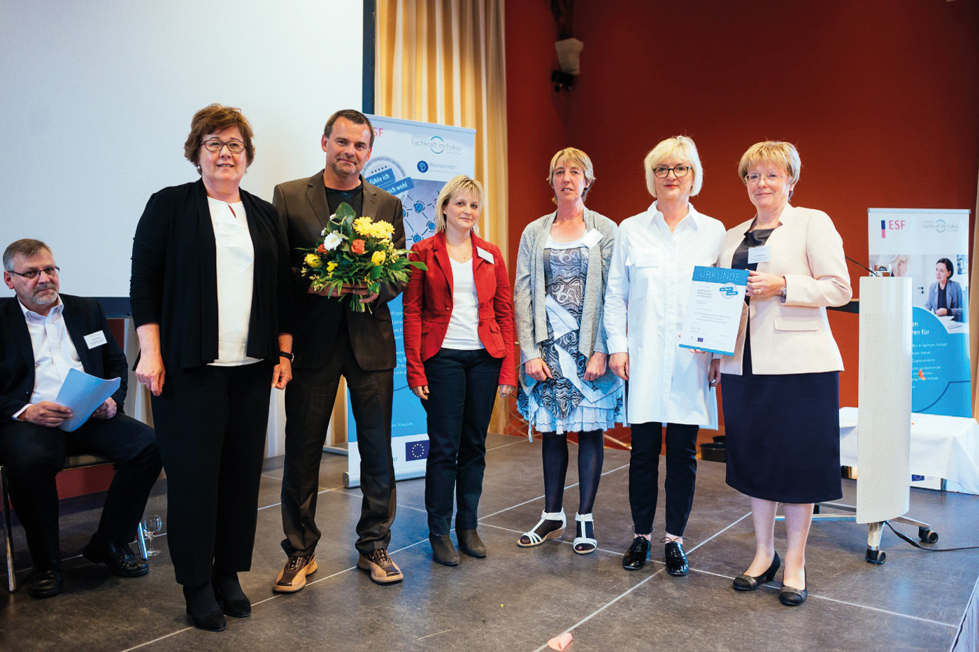 Links steht die Ministerin für Arbeit, Soziales und Integration Sachsen-Anhalt. Rechts neben ihr steht ein Mitarbeiter und 4 Mitarbeiterinnen der habils. Sie haben eine Urkunde und Blumen in er Hand. Alle schauen in die Kamera.