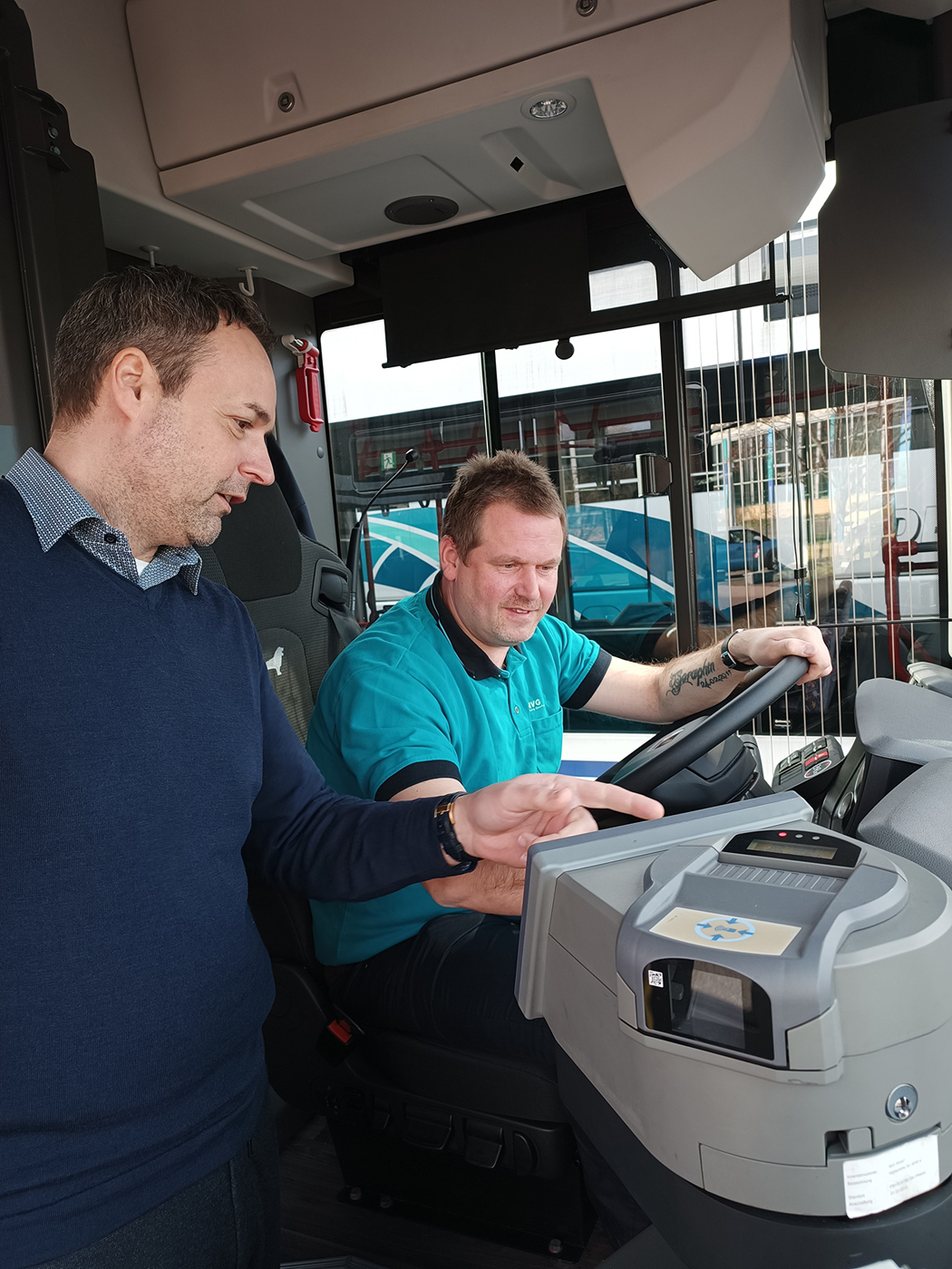 Ein Mann sitzt am Steuer eines Busses, ein zweiter erklärt ihm etwas an der Technik