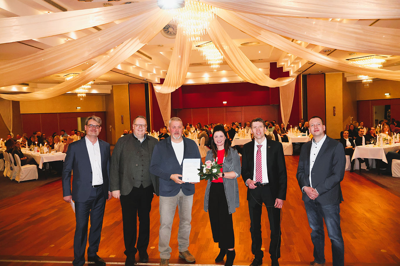 sechs Personen stehen in einem Festsaal auf der Bühne, im Hintergrund sitzen viele Menschen an festlich gedeckten Tischen.
