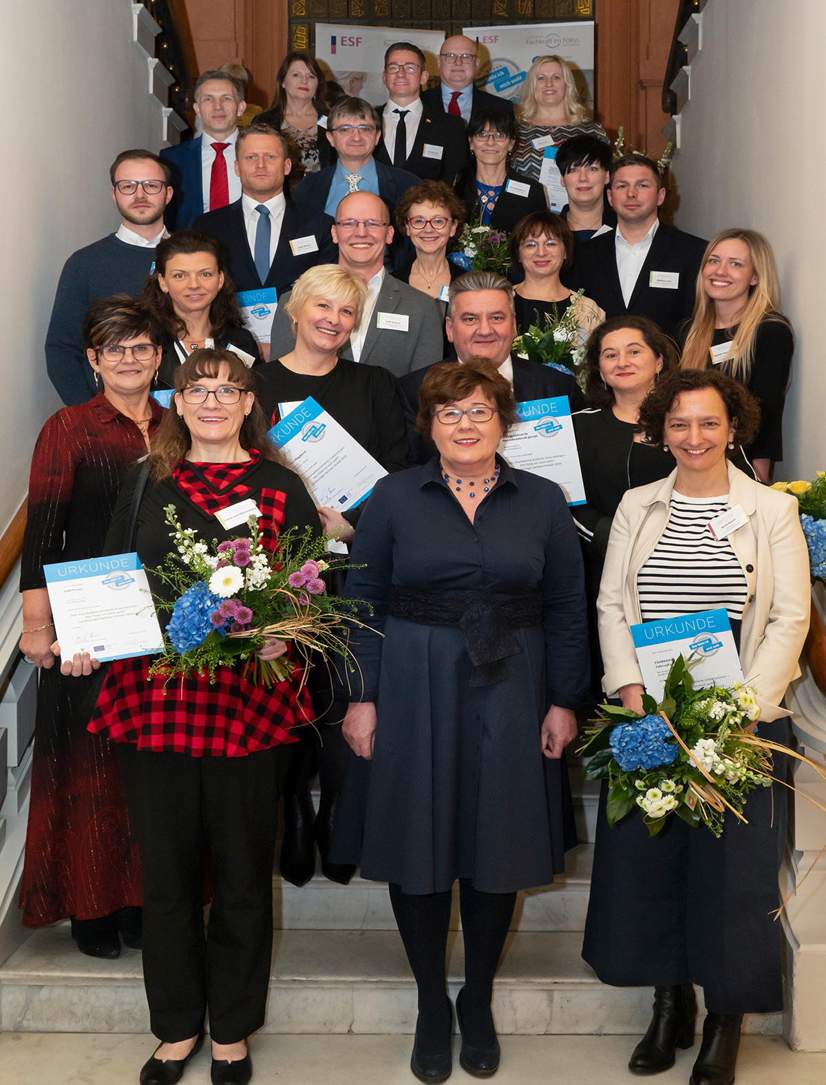 Frauen und Männer stehen auf einer Treppe, festlich gekleidet mit Blumen und Urkunden. Sie lachen in die Kamera